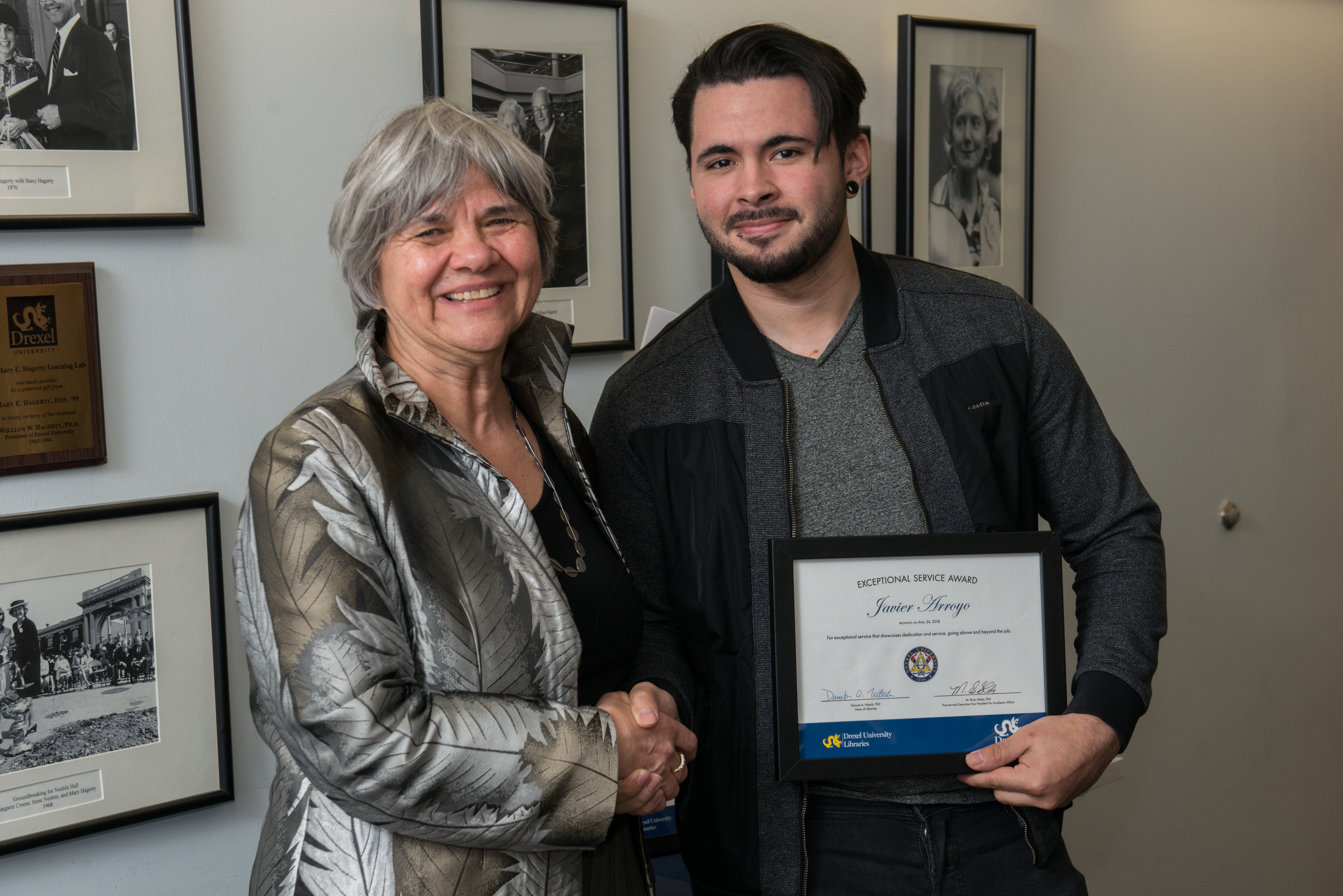 A woman and a man smile for the camera. They are shaking hands and the man holds a framed award.